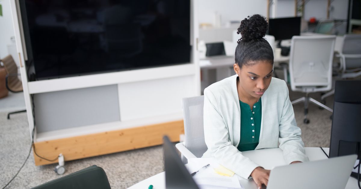 Focused woman using a laptop in a modern office setting, wearing a white blazer, working diligently.