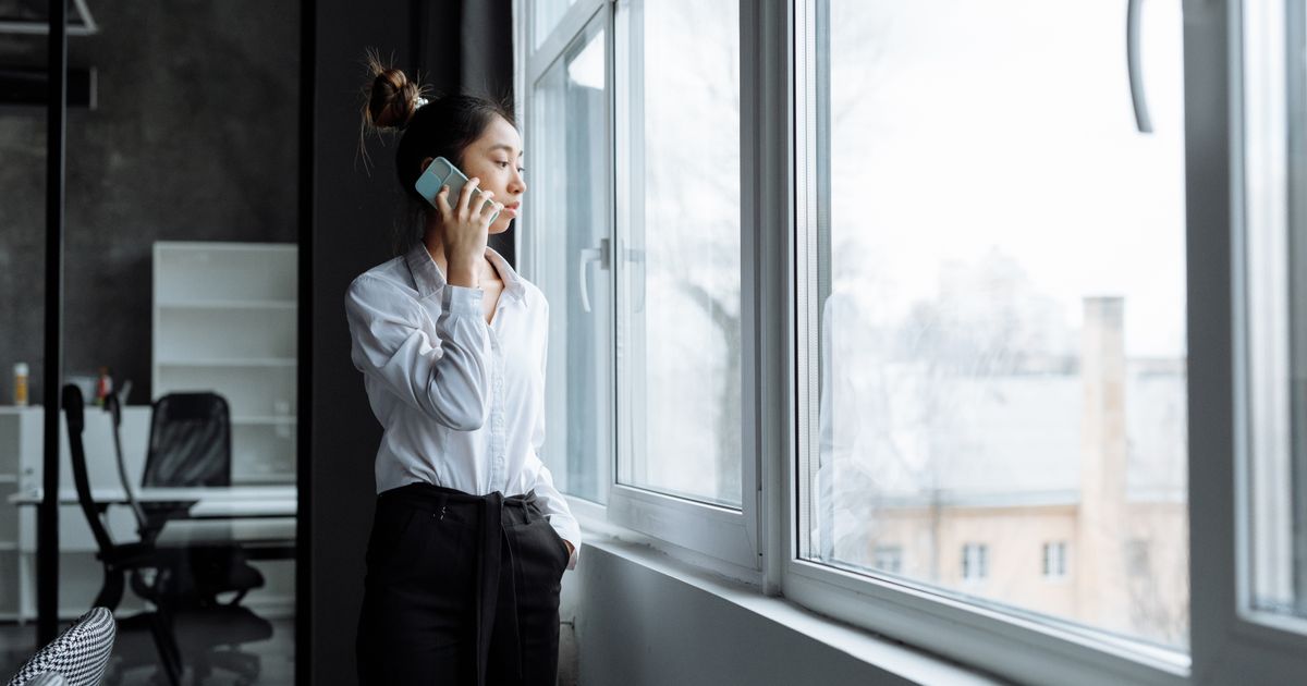 Asian woman in office clothes making a phone call while gazing outside a large window.